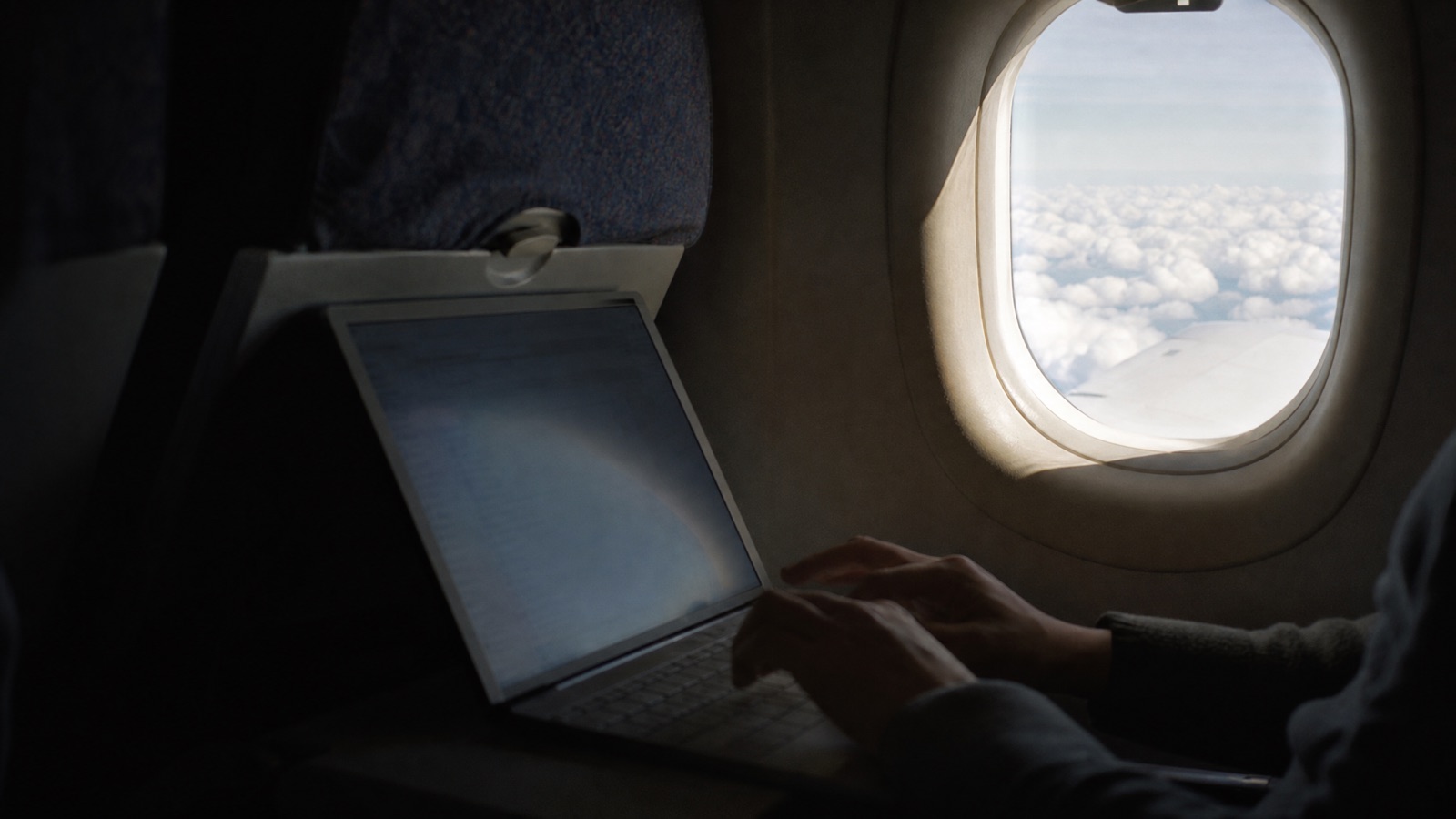 Passenger working on a laptop next to an aircraft window during daytime cruise, soft natural light, muted editorial tones.