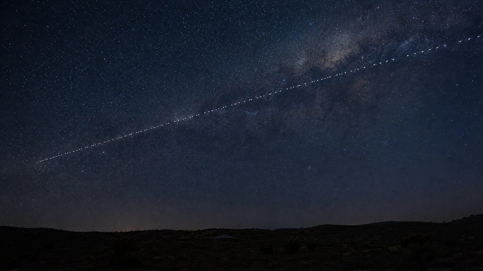 Long-exposure night-sky photograph of a faint Starlink satellite train tracing a diagonal line across a star-filled sky above a dark Western Australian ridgeline.