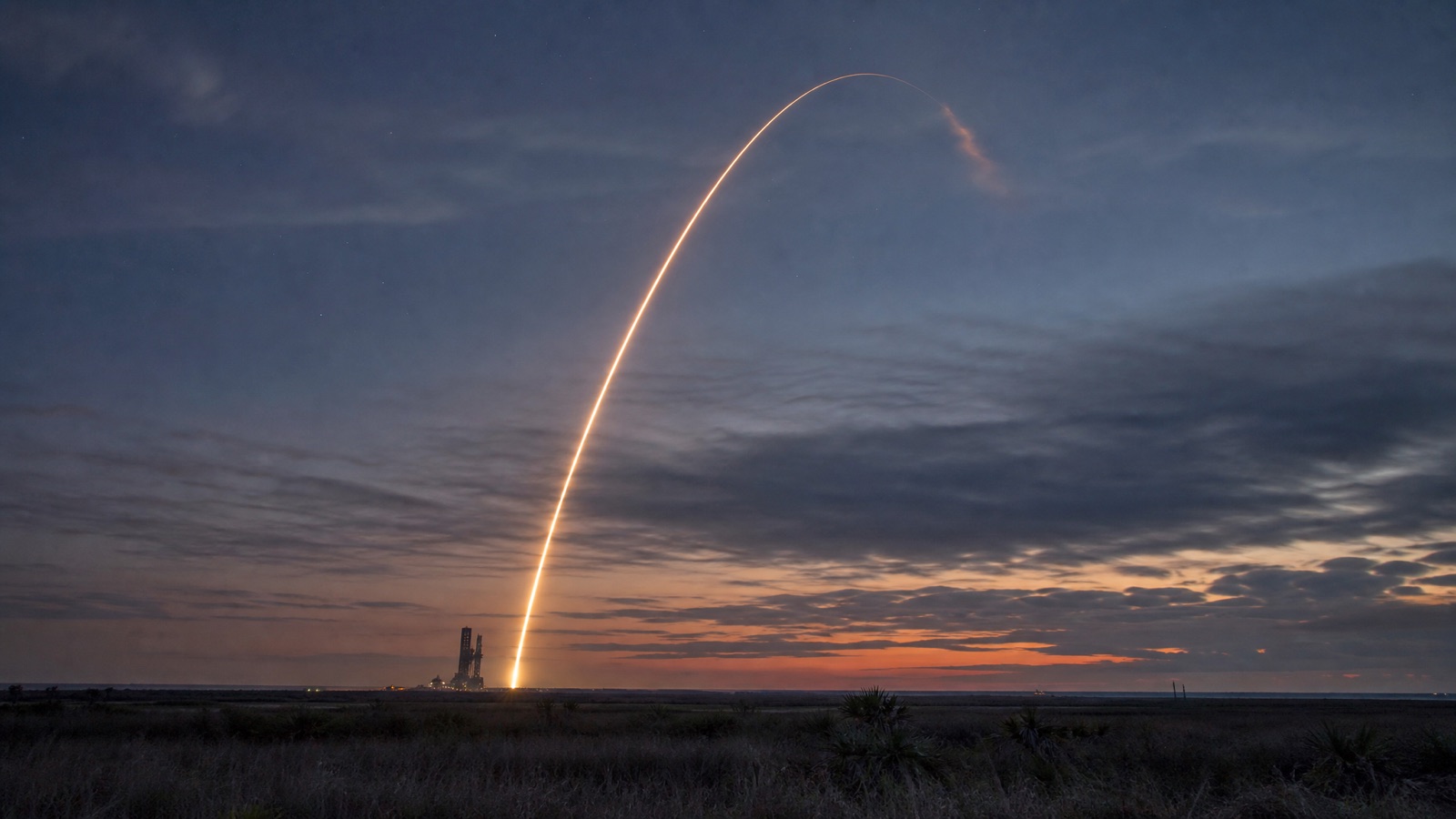 Long-exposure photo of a Starship-style rocket launch trail rising into a twilight sky over the South Texas coast